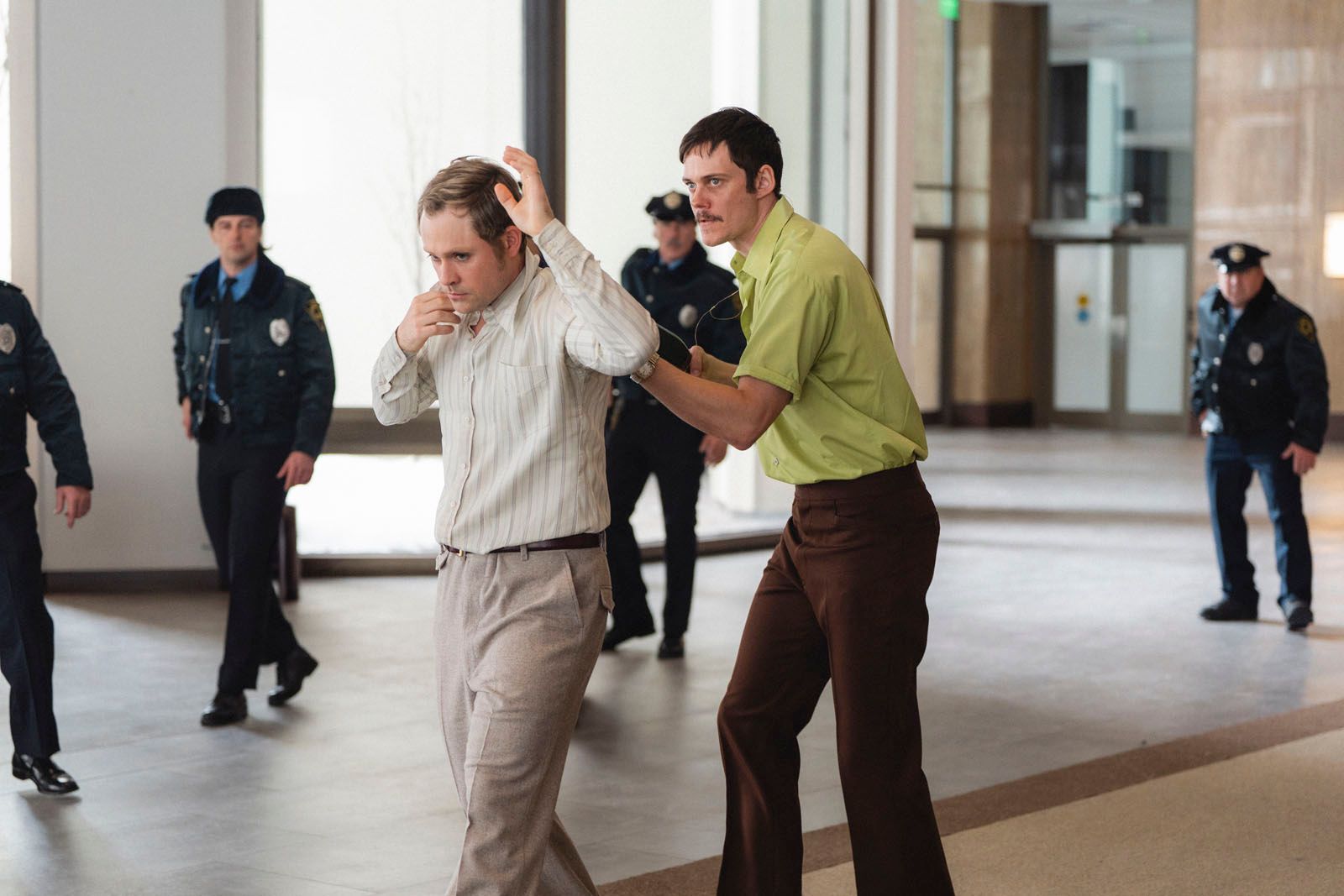 A scene in a modern building where a man in a white striped shirt is being escorted by another man in a green shirt, while several police officers watch in the background.